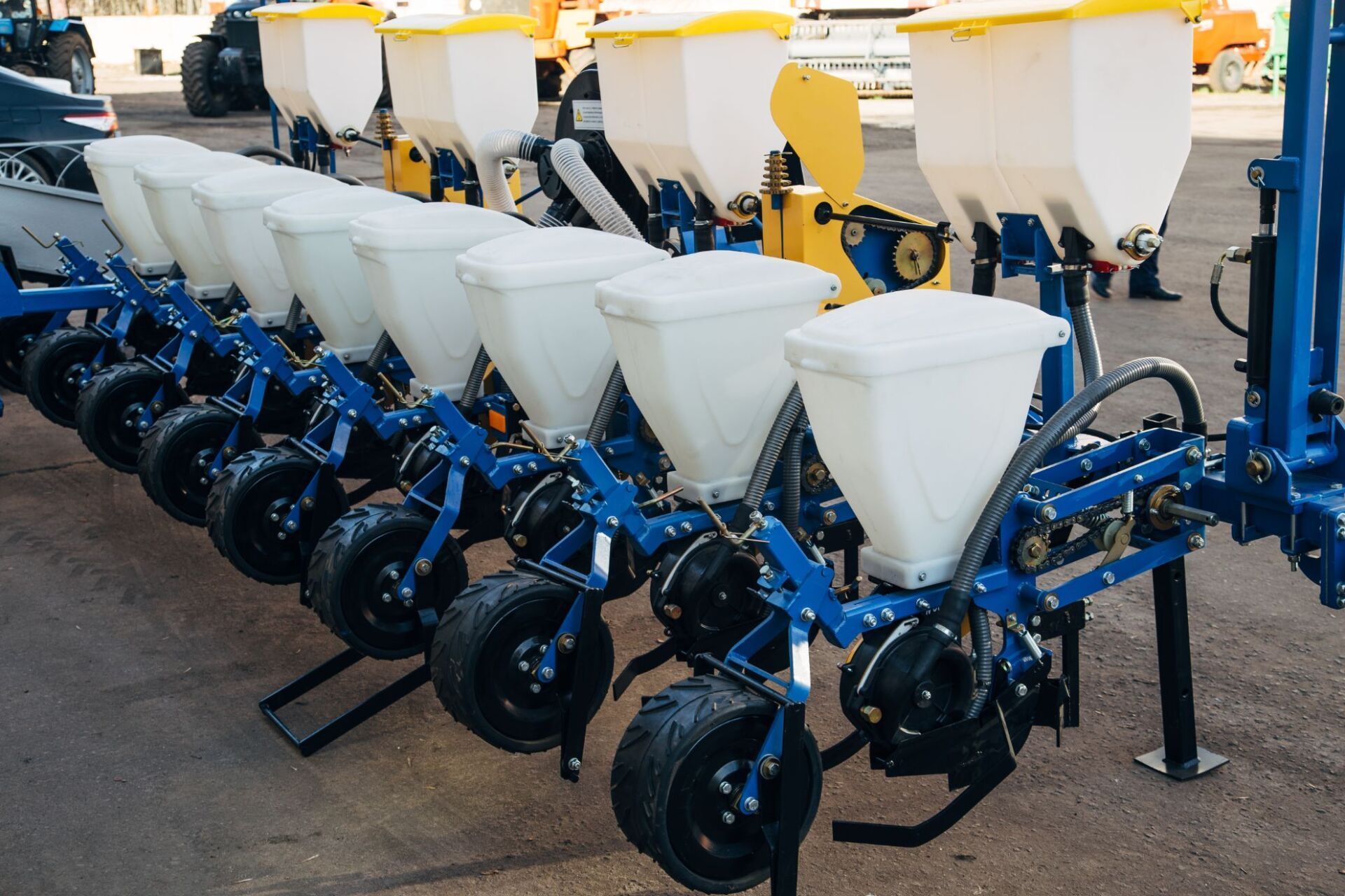 Una seminatrice agricola a più file con tramogge bianche e telai metallici blu parcheggiata su un piazzale asfaltato all'aperto.