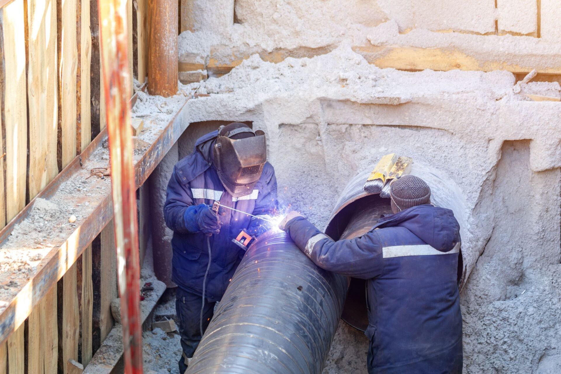 Due operai con indosso indumenti protettivi blu scuro saldano un grande tubo metallico all'interno di una profonda trincea di costruzione con pareti rinforzate.