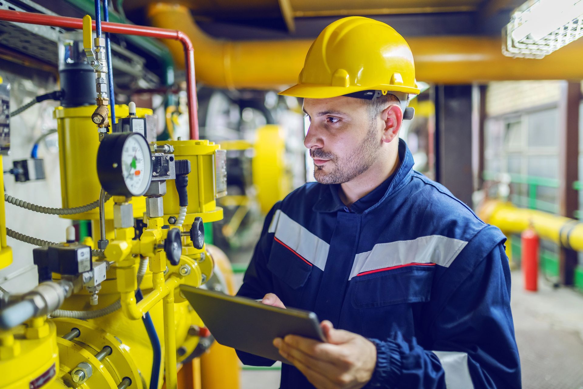 Un tecnico in uniforme blu e casco giallo tiene in mano un tablet mentre ispeziona macchinari industriali per il gas.