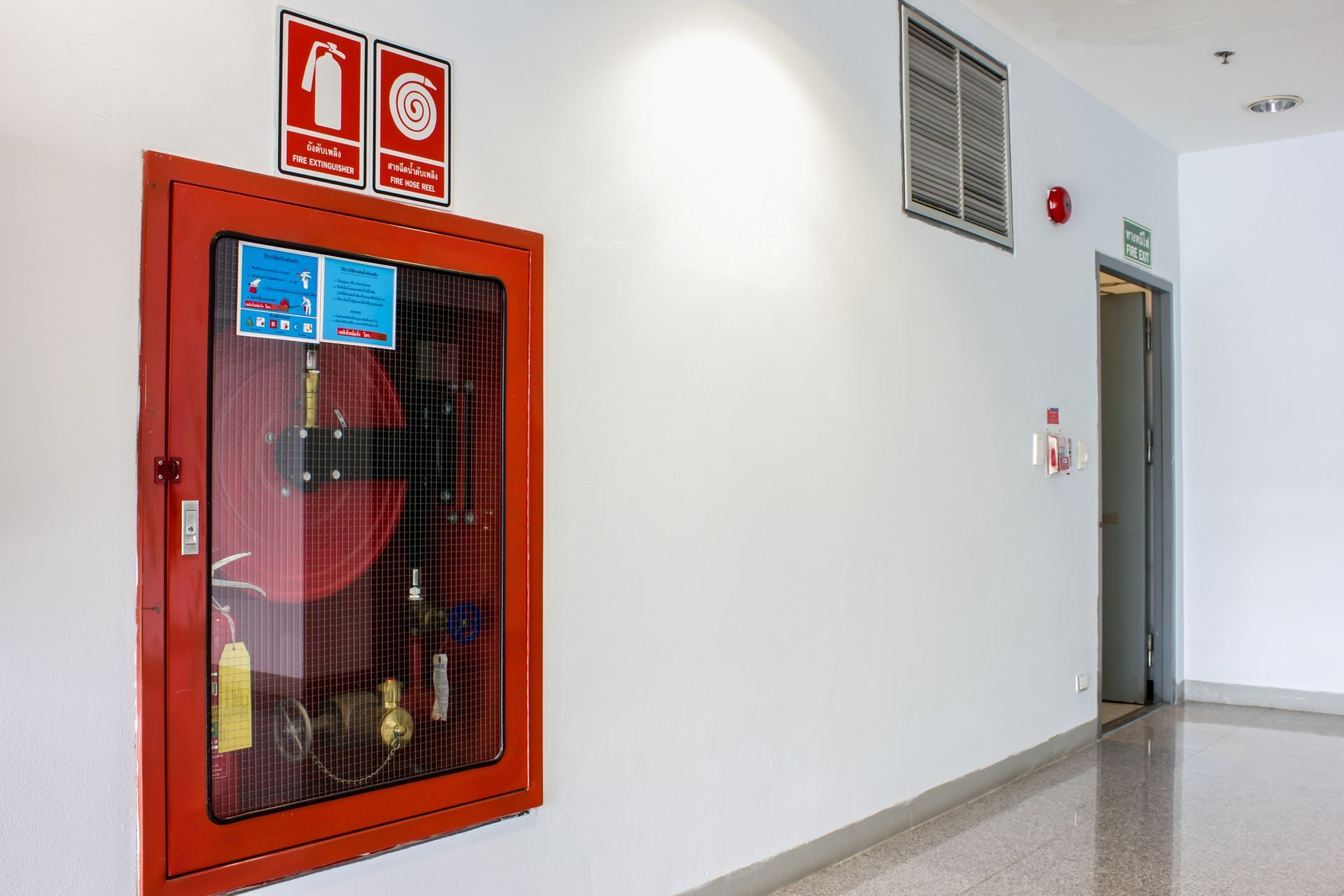 Un armadietto rosso per manichette antincendio montato su una parete bianca, con cartelli di sicurezza antincendio sopra, in un luminoso e moderno corridoio di un edificio.