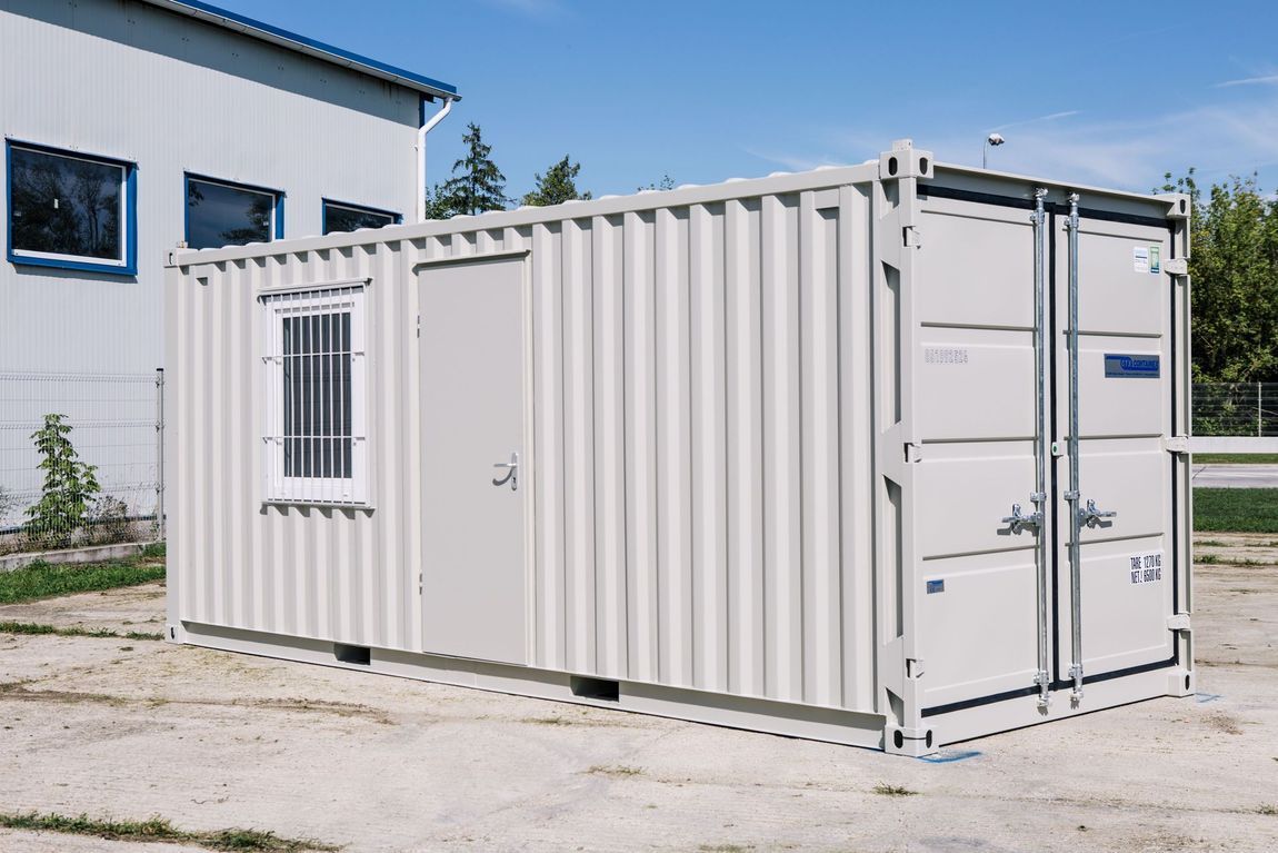 Un ufficio ricavato da un container color crema, con una sola porta e finestra, situato su un terreno ghiaioso.