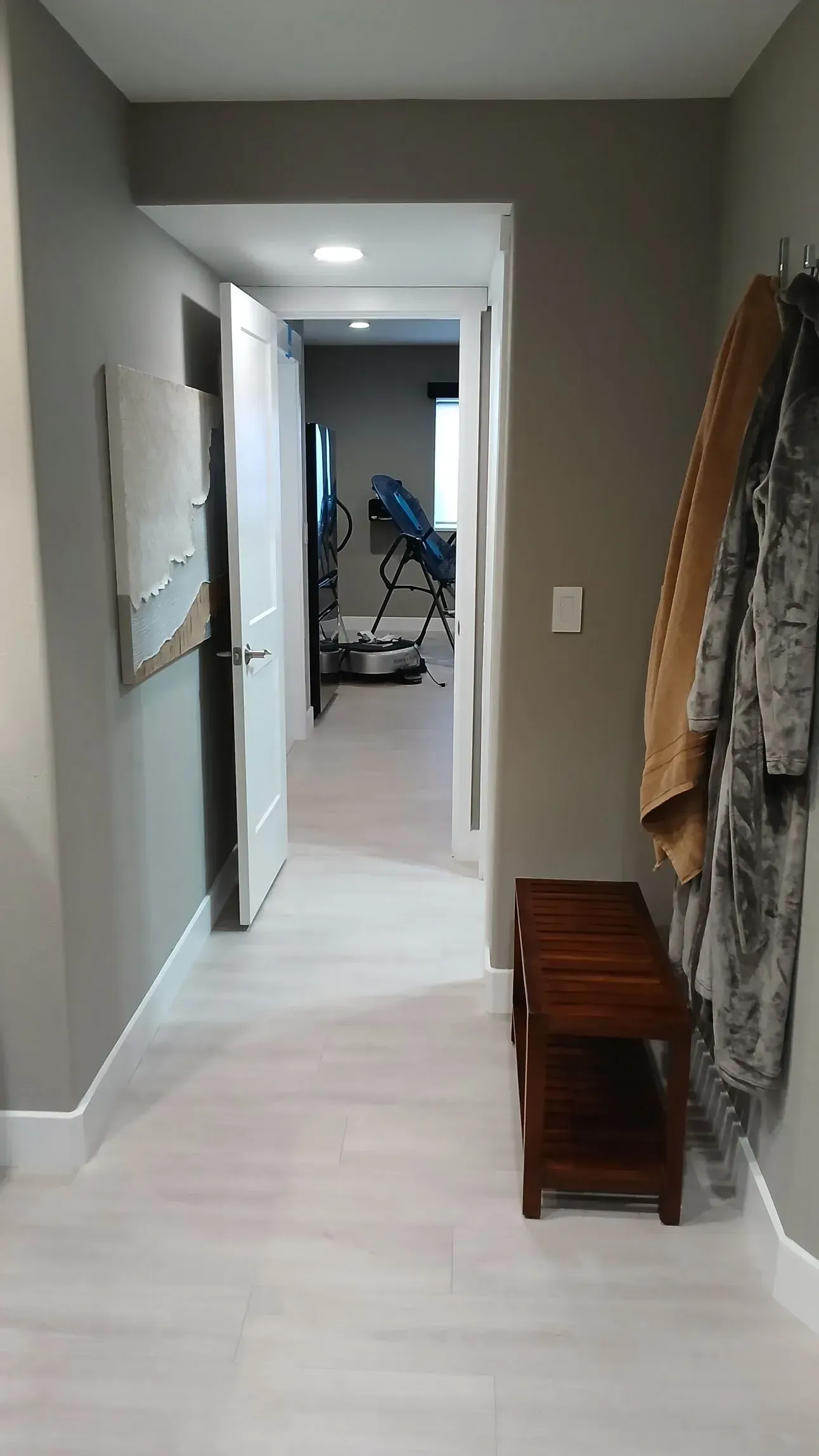 Hallway with light wood floors, gray walls, open door to room with blue chair, bench.
