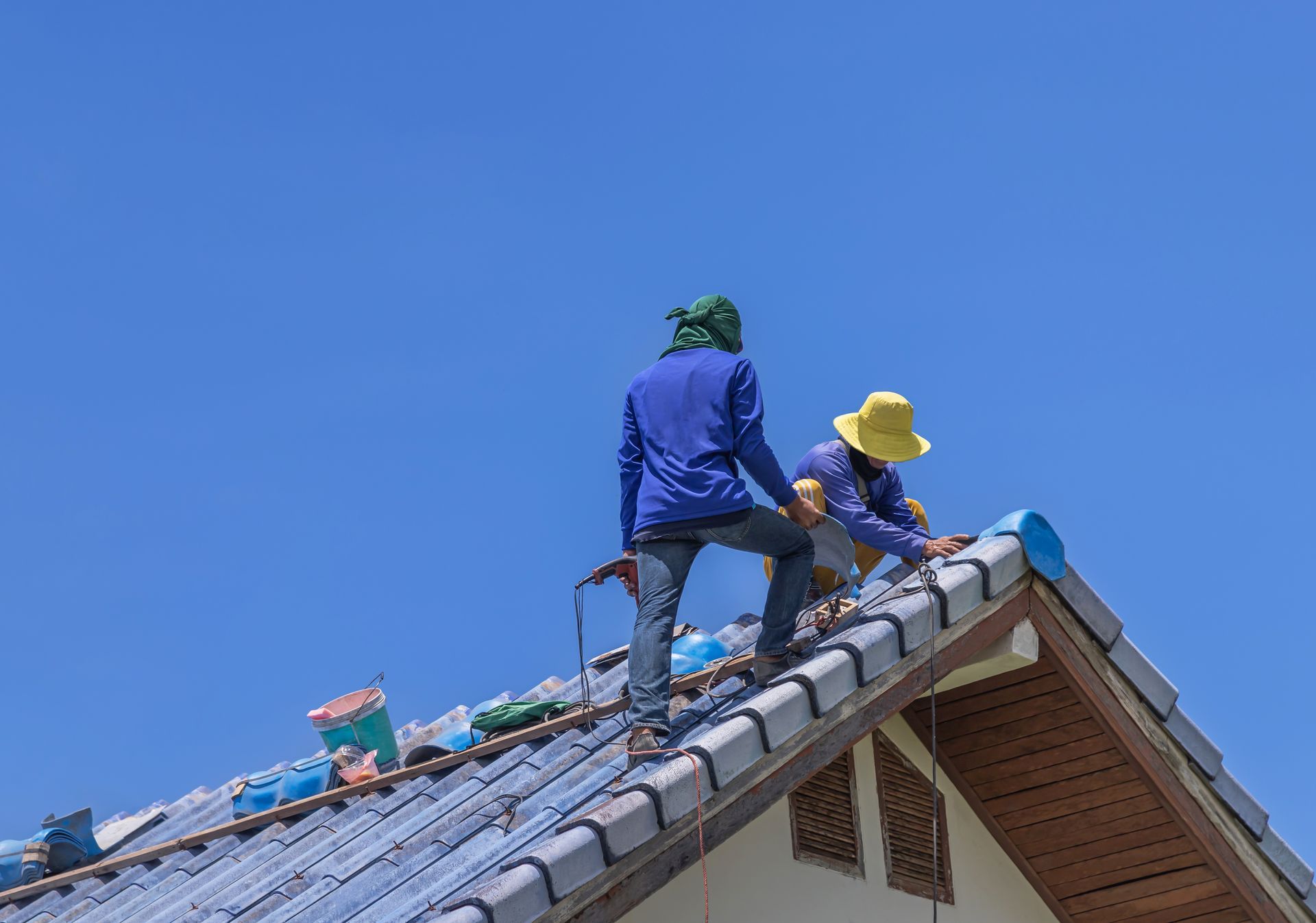 Two roofers on a blue-tiled roof under a clear, blue sky. One is drilling, the other is working.