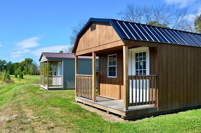 Two small wooden cabins with porches on a grassy rural lot under a blue sky