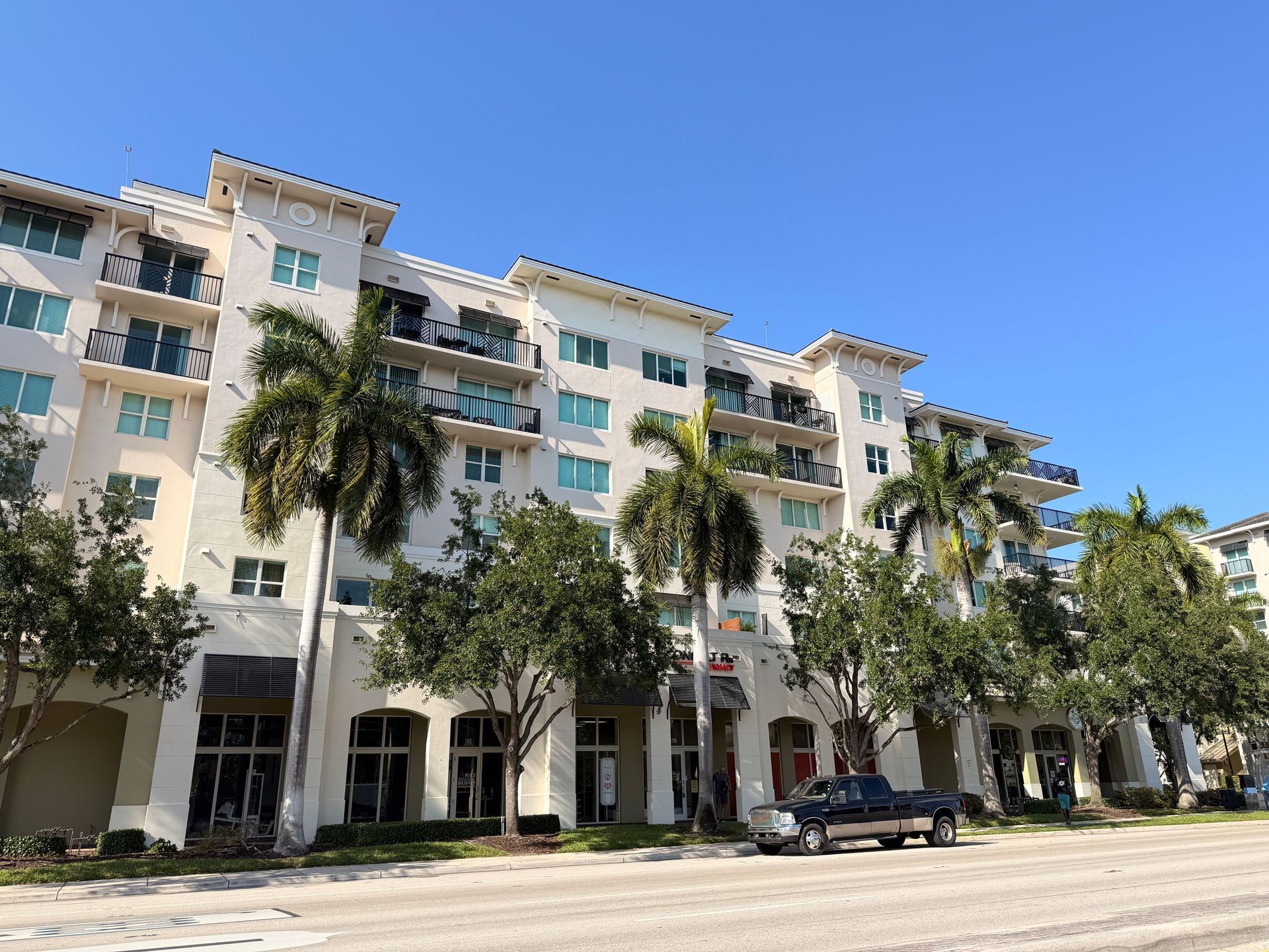 White beachfront apartment building with palm trees and a parked car under a clear blue sky