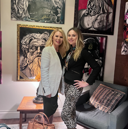 Two women posing in an art-filled living room beside a gray couch and patterned pillows.