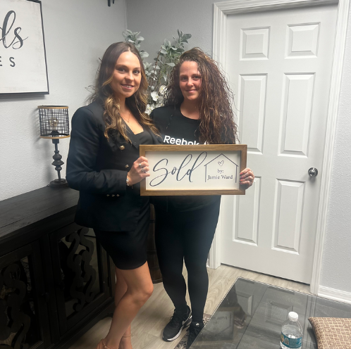 Two women hold a framed “Sold” sign in an office lobby.