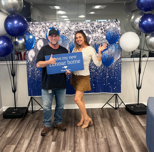 Two people posing at a real estate event, holding a “Let me help you find a home” sign with blue balloons