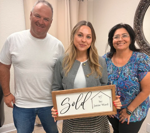 Three people smiling indoors, holding a framed “Sold” sign.