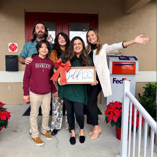 Five people smiling on a porch, holding a framed sign, with holiday decorations and a FedEx box behind them