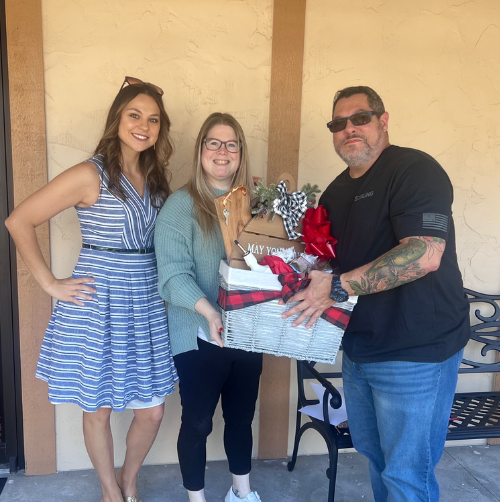 Three people posing indoors, holding a gift basket and wrapped gifts, smiling at the camera.