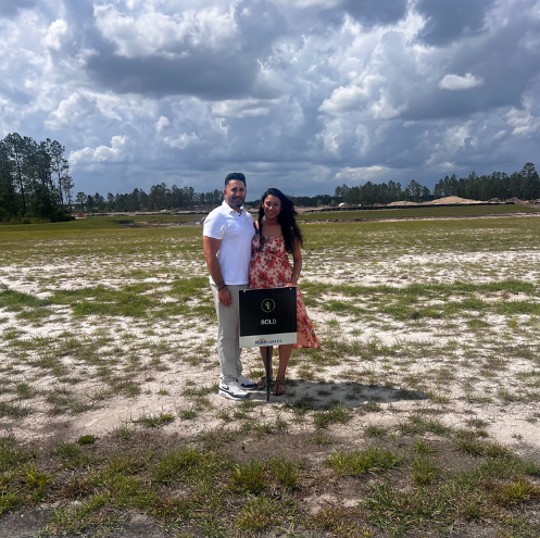 Two people standing in a grassy field under a cloudy sky, holding a sign.