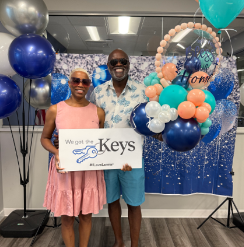 Two smiling people holding a “We got the keys” sign at a balloon-decorated celebration.