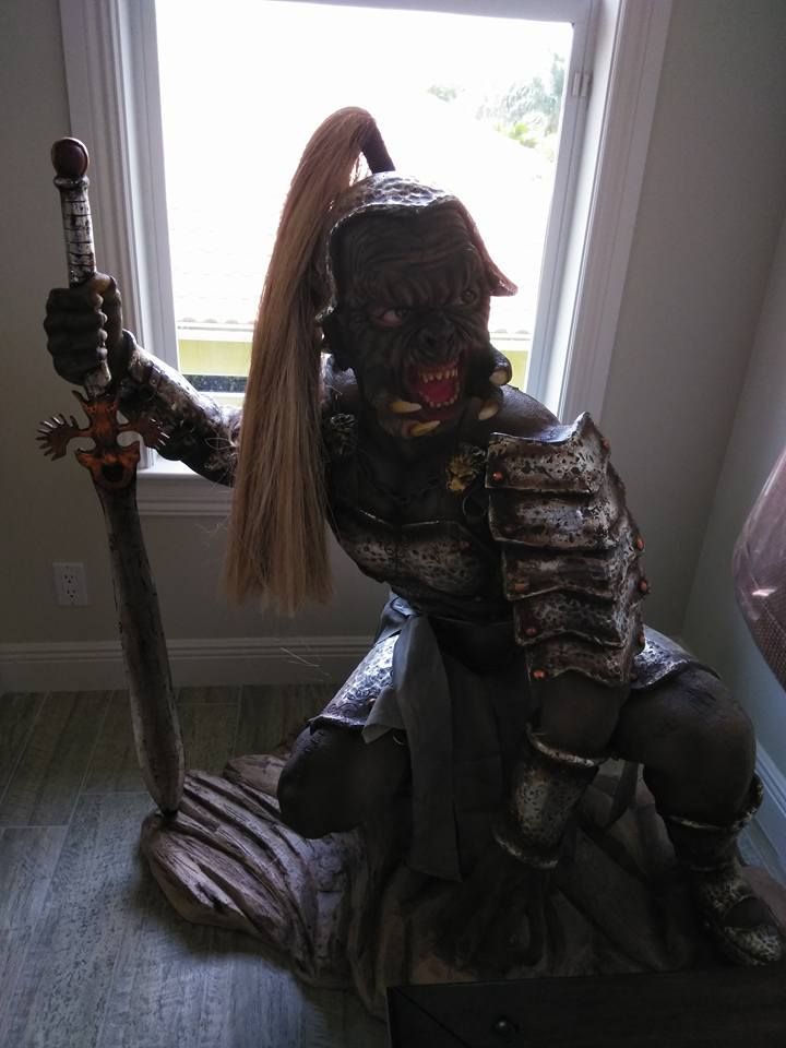 A dark, armored orc statue kneels indoors, holding a large sword and featuring a long, light-colored ponytail.