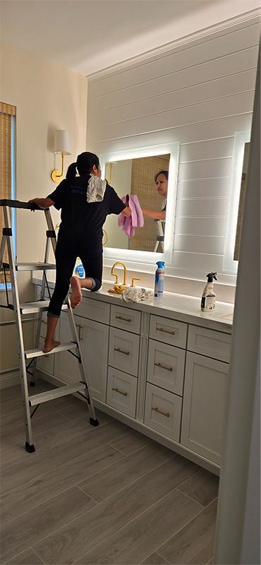 A person on a ladder cleans a bathroom mirror above a white vanity cabinet with gold hardware.
