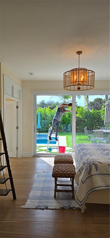 A person stands on a small step ladder inside a bedroom, cleaning the interior of a large glass sliding door to a patio.