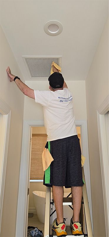 A person stands on a ladder in a hallway, using a yellow cloth to clean a square ceiling vent near a recessed light.
