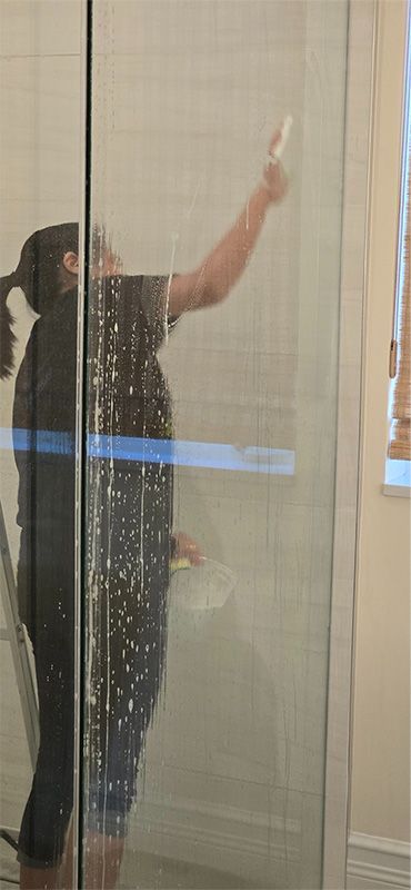 A person reaches up to clean a tall glass panel or door using a squeegee.