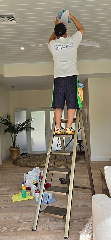 A person in a white shirt and dark shorts stands on a ladder, wiping down a white ceiling fan in a bright living room.
