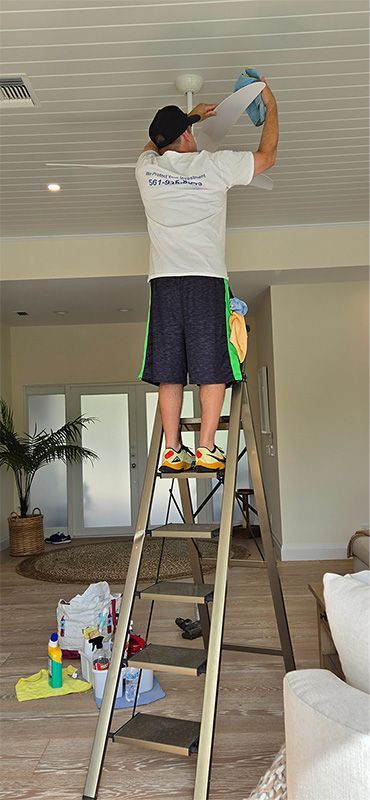 A person stands on a ladder cleaning a ceiling fan inside a home.