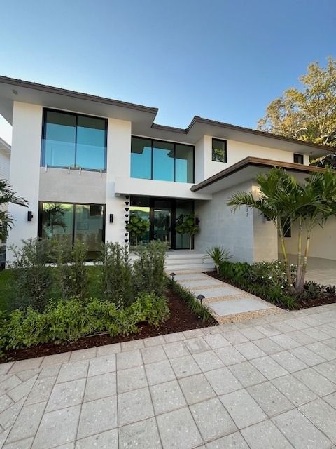A modern two-story white house with large glass windows, a paved walkway, and landscaping under a clear blue sky.