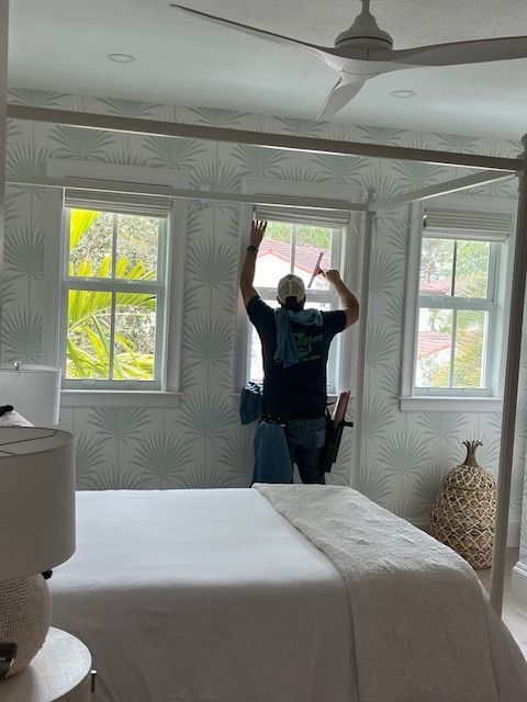 A person stands on a bed, installing a curtain rod above a bedroom window against a patterned, light-colored wallpaper.