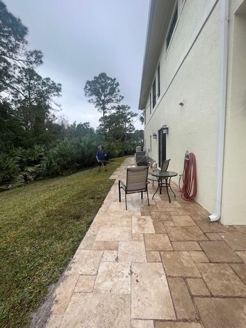 A stone patio adjacent to a light-colored house wall, featuring a small table and chair, with a lawn and trees nearby.