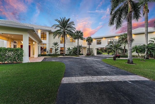 A large, beige mansion with a paved circular driveway and palm trees under a dramatic, colorful sunset sky.