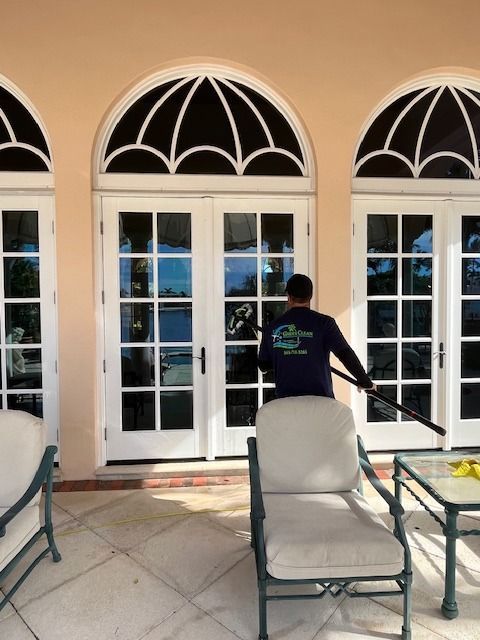 A person wearing a dark long-sleeved shirt cleans a large glass double door on a patio with a pole-mounted squeegee.