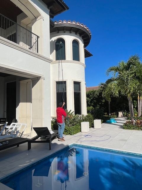 A person in a red shirt uses a long telescopic pole to clean the windows of a white, multi-story house near a pool.