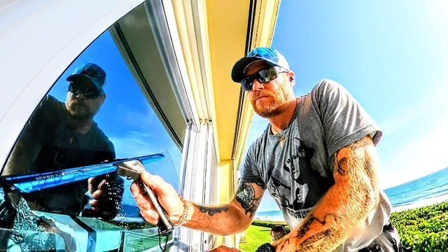 A person with tattoos wears sunglasses and a cap while using a squeegee to clean a window near the coast.