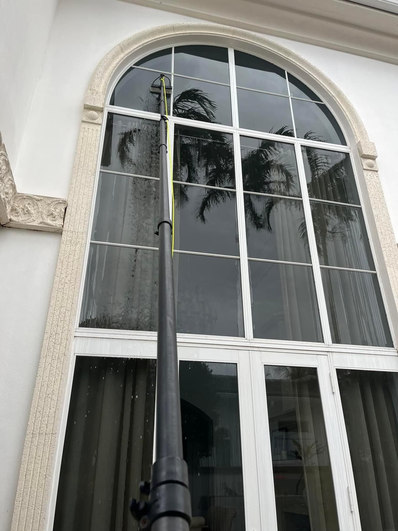 A long, black extension pole reaches up to clean the glass of a large, white-framed arched window on a building.