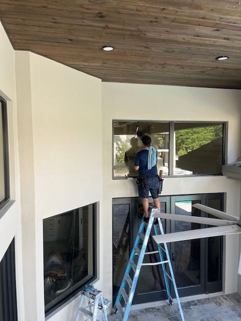 A person stands on a ladder cleaning a high window on the exterior of a modern, cream-colored home with a wood ceiling.