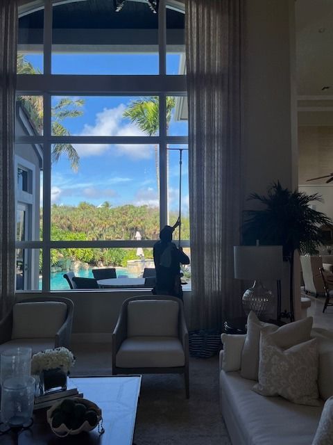 A professional window cleaner uses an extended pole to wash the tall, arched windows of a sunlit living room.