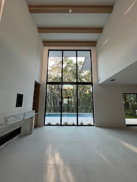 A modern room with high ceilings, wooden beams, a marble fireplace, and floor-to-ceiling windows overlooking a pool.
