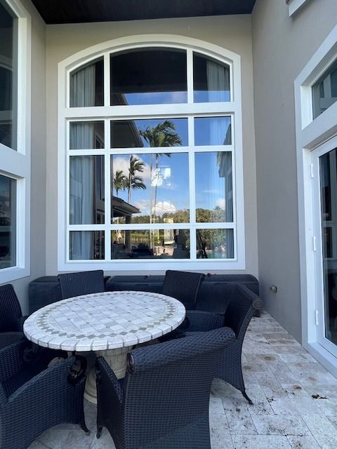 A patio with a round tiled table and dark wicker chairs set against a large arched window on a stone-tiled floor.