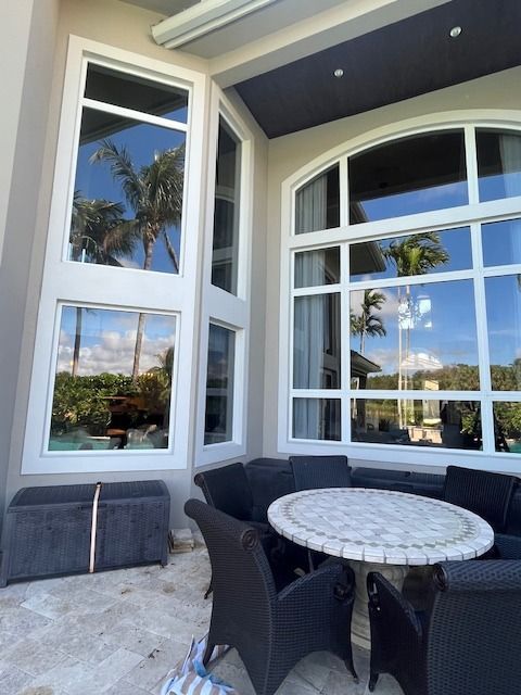 Patio with a round mosaic table and dark wicker chairs beside tall, white-framed windows reflecting palm trees.