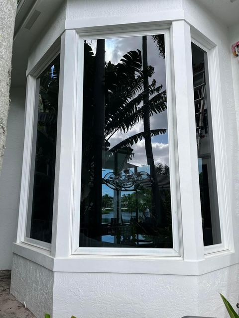 A white bay window with tinted glass reflecting palm trees and the sky, viewed from an exterior walkway.