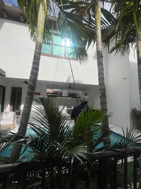 A person standing by a backyard pool uses a long pole to clean the exterior second-story windows of a white house.