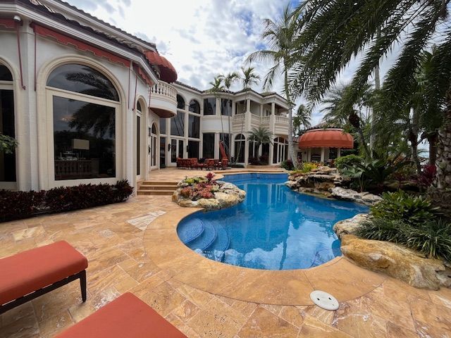 A large luxury home with a swimming pool, stone patio, and palm trees under a cloudy sky.