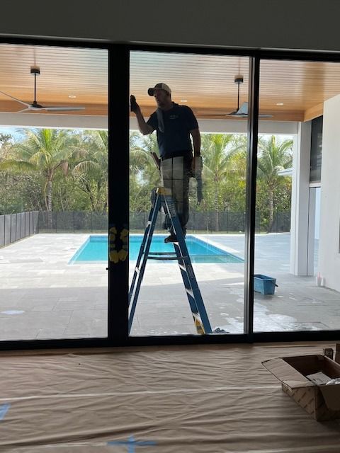 A person on a ladder cleans a large floor-to-ceiling glass door, looking out onto a backyard with a swimming pool.