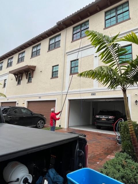 A person in a red jacket uses a long pole to clean the windows of a multi-story, beige townhouse complex.