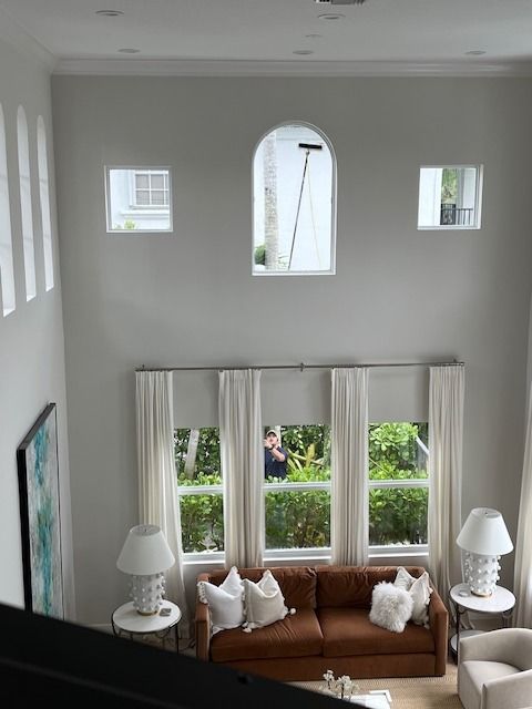 A high-ceilinged room with a brown sofa, white curtains, lamps, and several windows, including one arched window.
