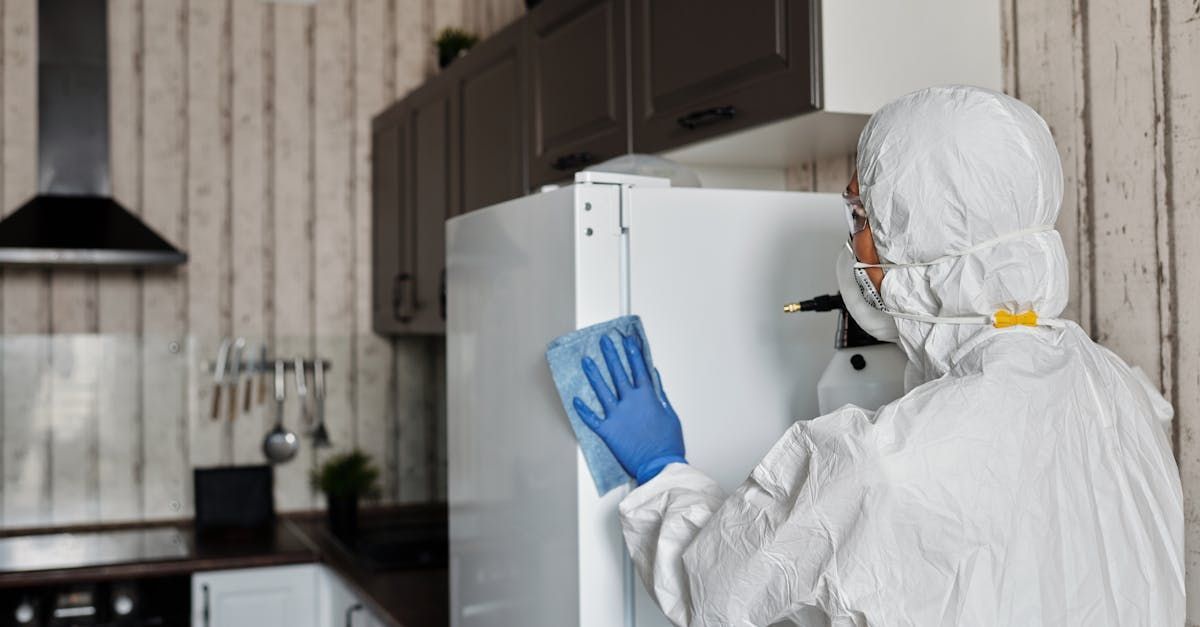 A person in a protective suit is cleaning a refrigerator in a kitchen.