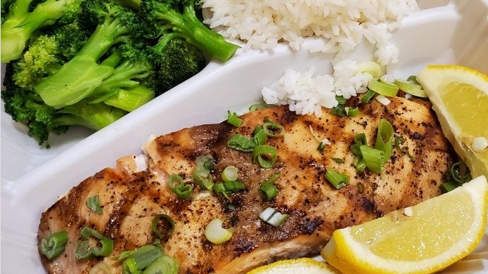A styrofoam container filled with grilled salmon , broccoli , rice and lemon slices.