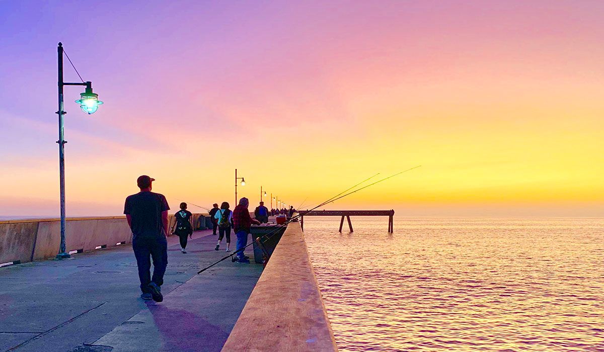A group of people are walking along a pier at sunset.