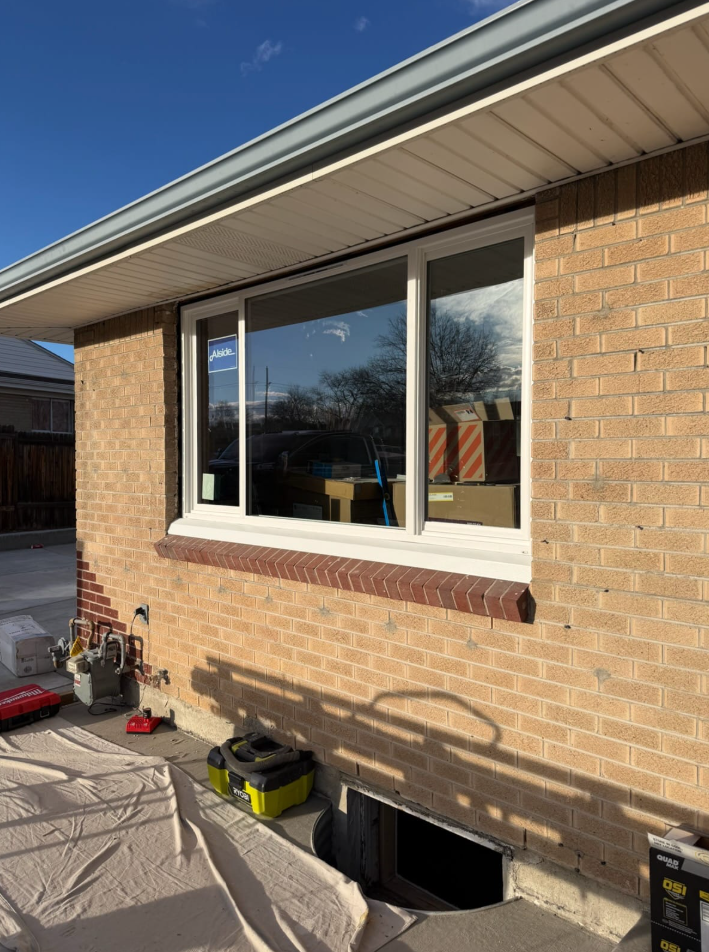 Brick house exterior with a large window, roof eave, and tools on a covered patio below the wall.