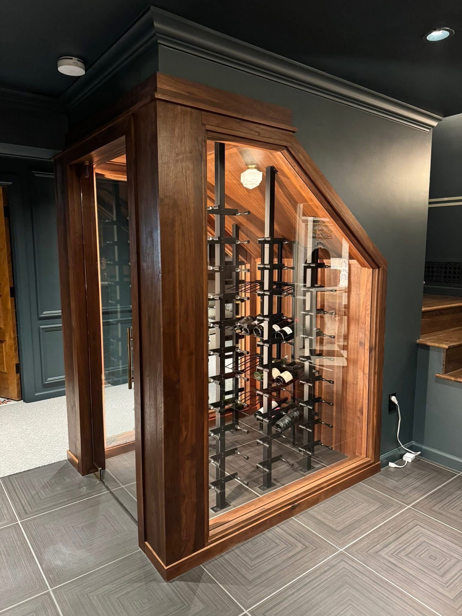 Wooden wine cellar with glass front and dark bottles, set in a gray-walled room with patterned flooring.