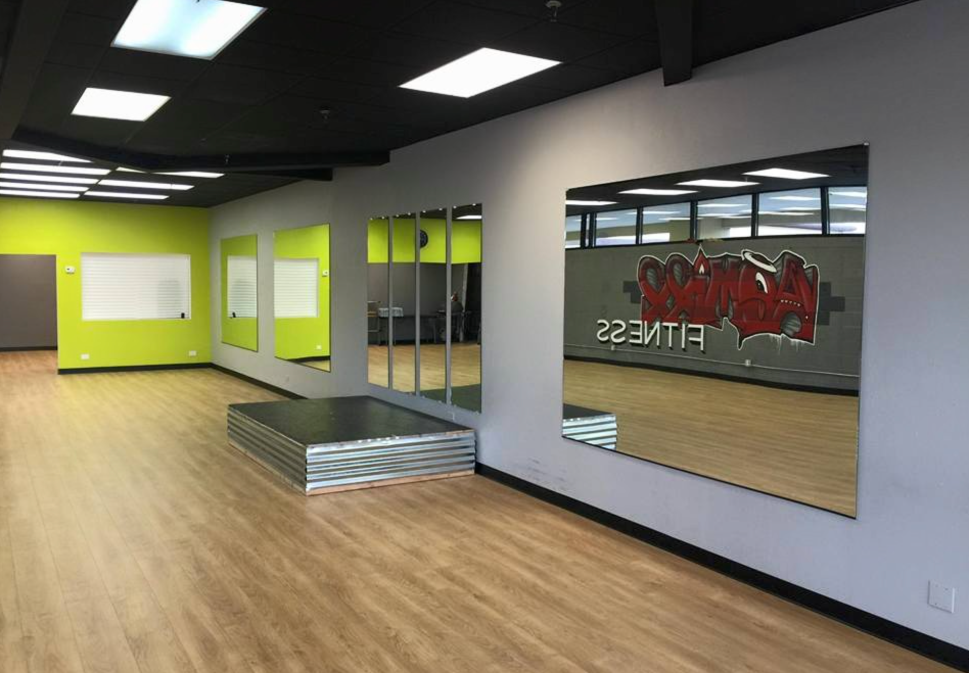 Empty gym with mirrors, stage, and logo on the wall. Floors are wood-like with green and gray walls.