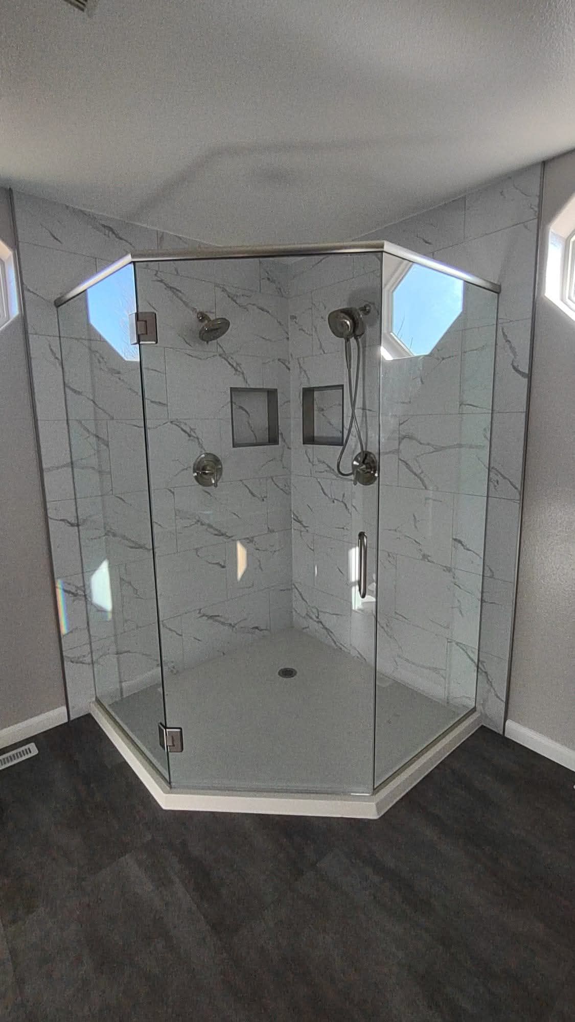 A modern, tiled shower with glass doors. Two shower heads and two niches are visible. Dark flooring.